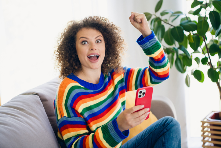 Portrait of pretty young woman raise fist hold smart phone wear sweater modern interior flat indoors