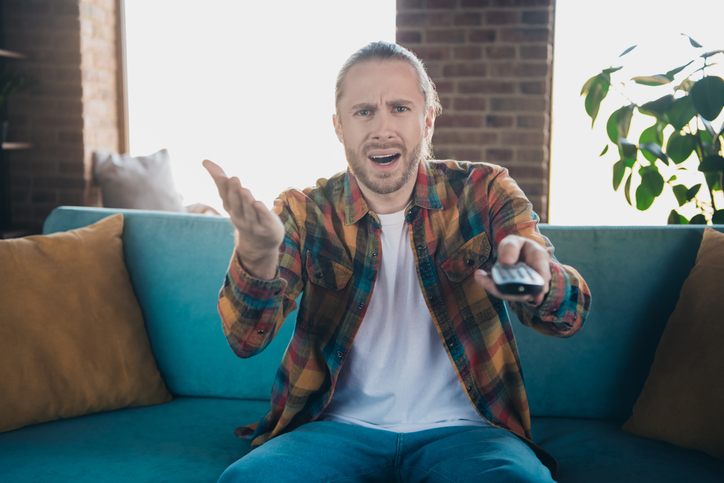 Young male watching TV on a weekend in a trendy loft living room wearing a plaid shirt, expressing surprised emotions