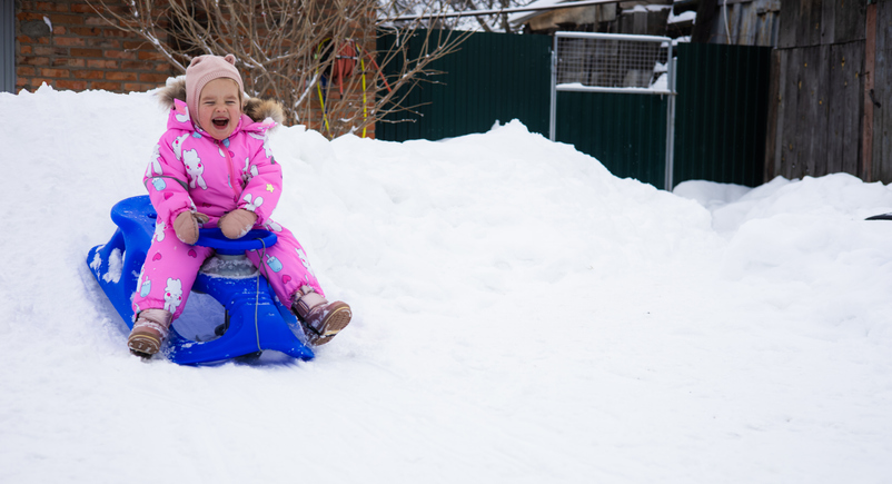 A child sleds down a snowy hill in the backyard, enjoying the winter. Copy Space. In a bright pink jumpsuit, a little girl sleds down a snow hill built by her parents. A fun winter day.