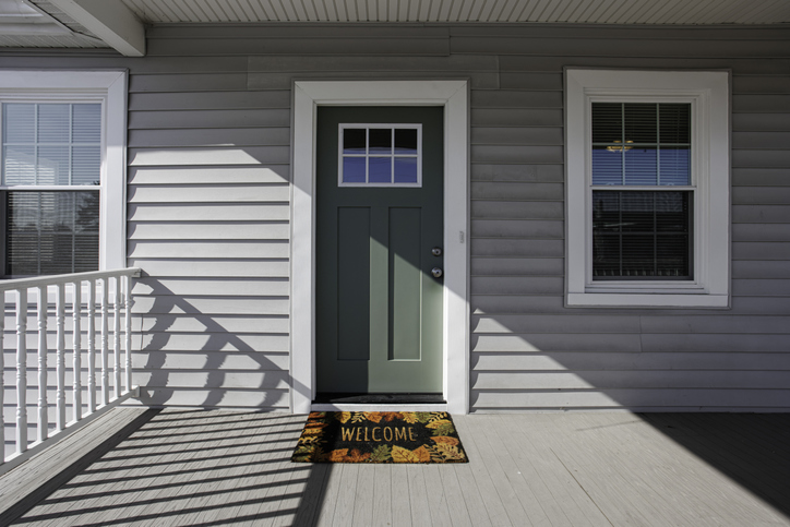 Green Door With Welcome Mat On Wooden Porch Area.