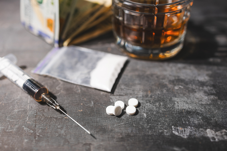 White narcotic pills, an alcoholic drink in a glass, syringe with a dose of heroin, cocaine powder in a transparent bag, and US dollar bills on a dark background. Concept of addiction and bad habits