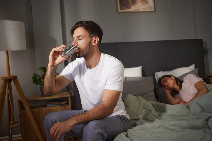 Man sitting on bed drinking water while woman sleeps peacefully