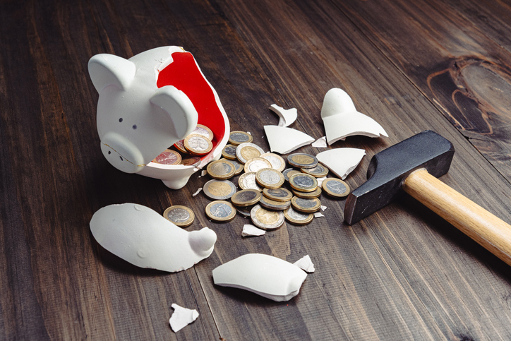 Broken white piggy bank with scattered Euro coins and hammer on wooden table, depicting financial crisis and savings loss