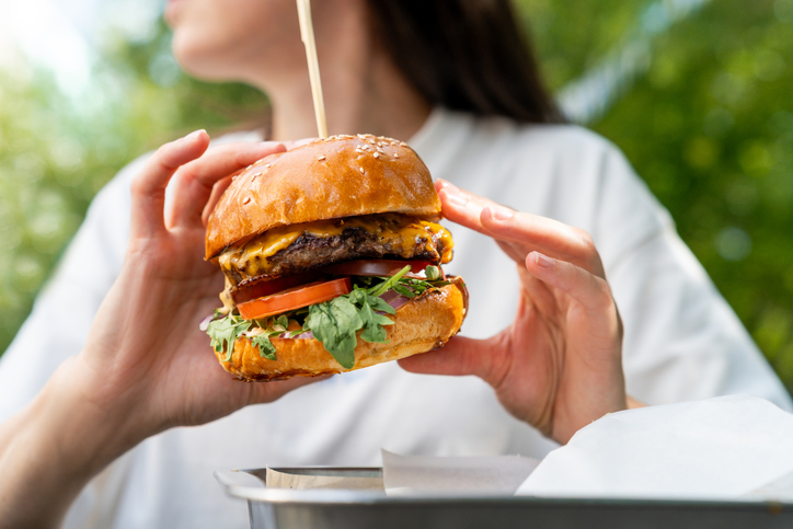 Close-up of a woman sitting at an outdoor garden table eating a juicy cheeseburger with salad and pickles
