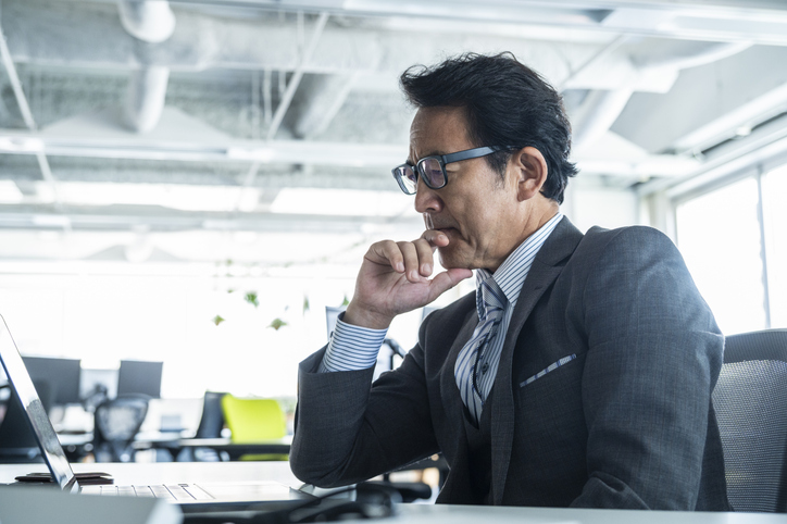 Thoughtful Japanese businessman looking at laptop in modern office space