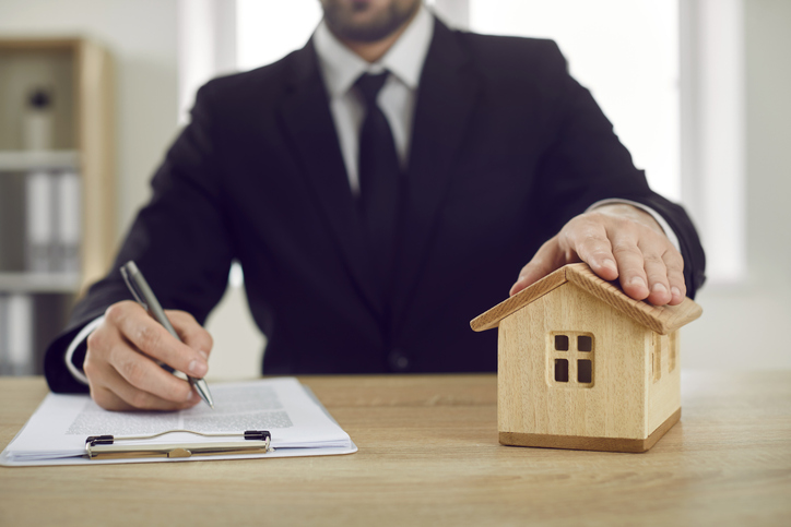 Man at real estate agent's office puts his hand on toy house and signs mortgage agreement