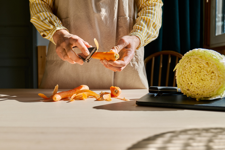 Hands of unrecognizable woman in apron peeling carrots using a food peeler.