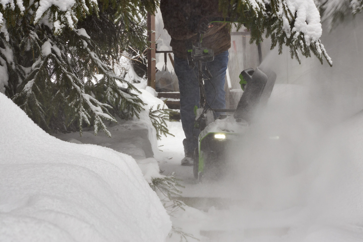 Man clearing a snow-covered garden path with a snow blower after heavy snowfall. Winter scene with deep snowbanks, evergreen trees, cold weather, seasonal work, and residential outdoor maintenance