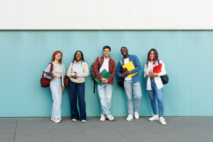 Happy multiethnic students standing against light blue wall, holding notebooks and backpacks