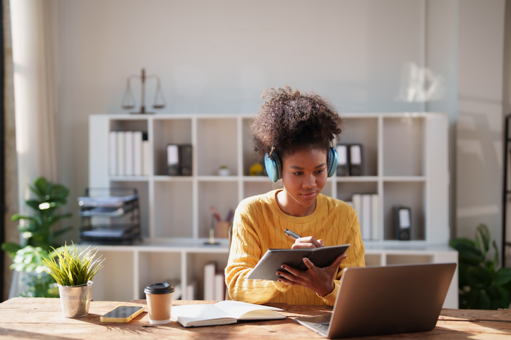 Young Black woman with headphones studying at a bright home office desk, focused on laptop and notebook as she takes notes and participates in online learning
