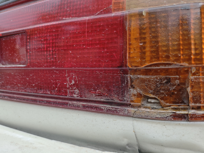 Close-up of a damaged, cracked, and weathered car taillight, showing signs of aging, neglect, and poor vehicle maintenance. Focus on red and amber sections