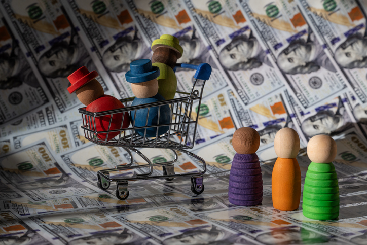 Small shopping cart with wooden figures and several peg dolls standing nearby, set against a backdrop of US dollar bills, illustrating family shopping and financial planning.