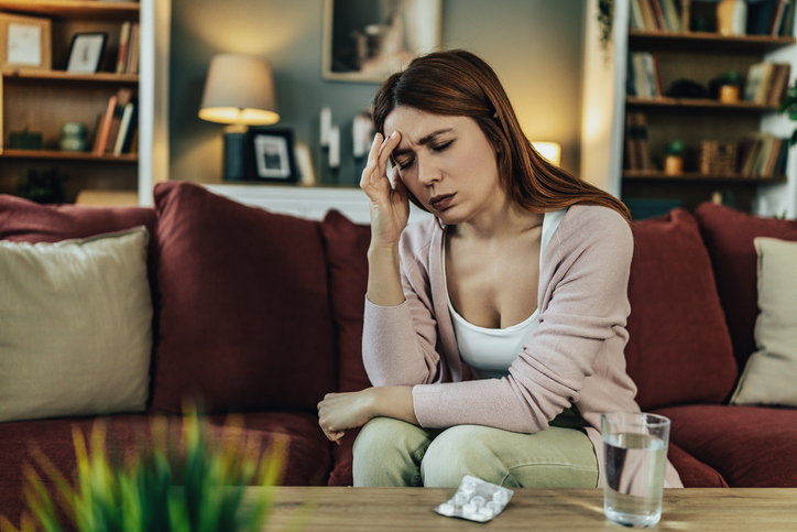 Stressed Woman Sits on Couch Holding Her Head as Medication and Water Glass Sit Nearby