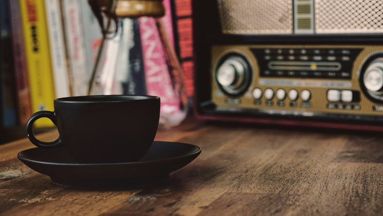 coffee brewing jug, vintage radio, a black coffee cup and saucer on a brown wooden table, with books in the background,