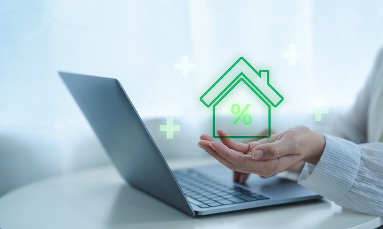 A person holds a glowing green house icon with a percentage symbol above a laptop, symbolizing online mortgage or real estate financing.