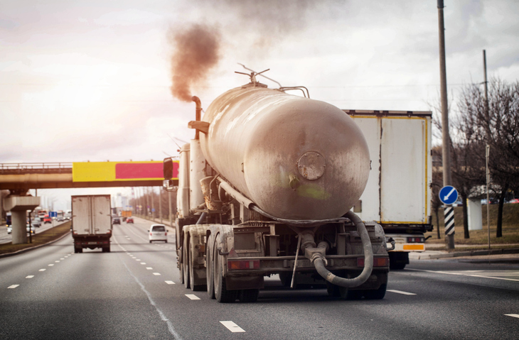 Tank truck travels on multi-lane highway, dark exhaust smoke billows from pipe, representing air pollution of heavy vehicle transportation. Copy space for text
