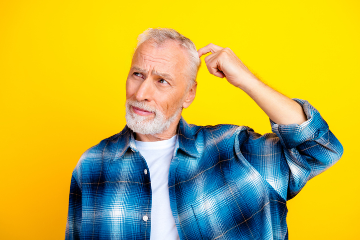 Confused senior man scratching his head and thinking in a bright studio portrait wearing a blue plaid shirt against a yellow background