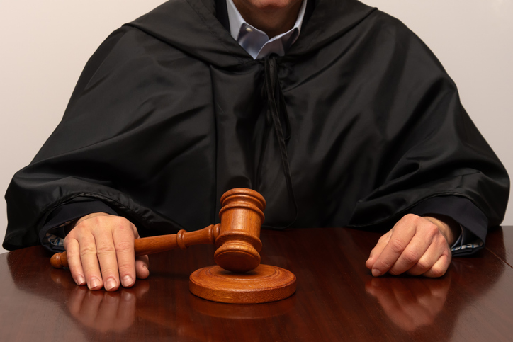 Judge in black robe holds wooden gavel at courtroom table during hearing.