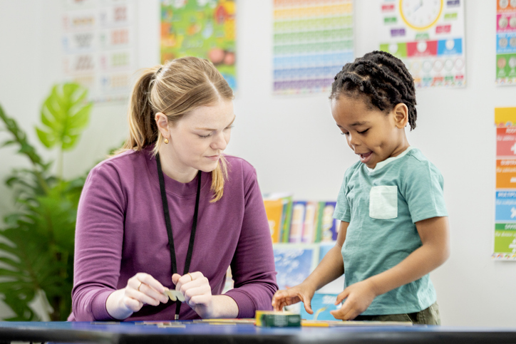 Female Teacher Helping Young Boy Student in Bright Elementary Classroom During Hands-On Learning