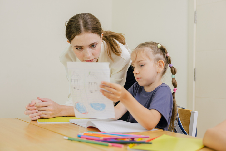 A young girl shows her drawing to a teacher at a desk with colored pencils and paper in a classroom setting. Back to school and education concept