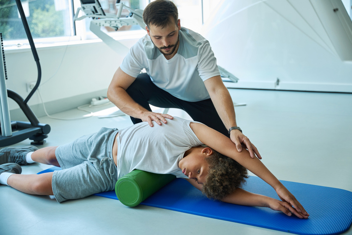 Man assisting boy with stretch using foam roller in gym