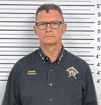 A middle-aged man wearing glasses and a sheriff's uniform stands in front of a lineup backdrop.