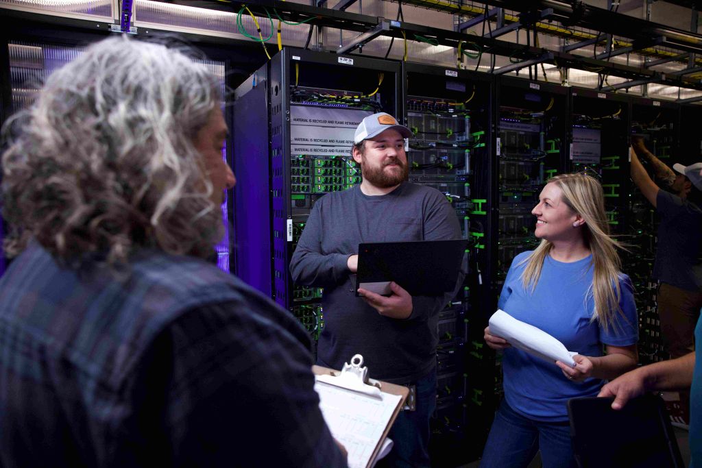 Three people in a data center, one holding a laptop and discussing with the others.