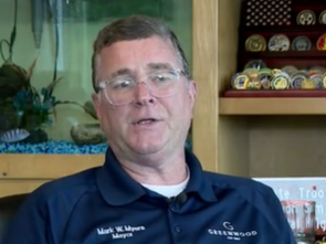 Middle-aged man wearing a navy blue shirt with a logo on the chest, standing in front of shelves displaying various items.
