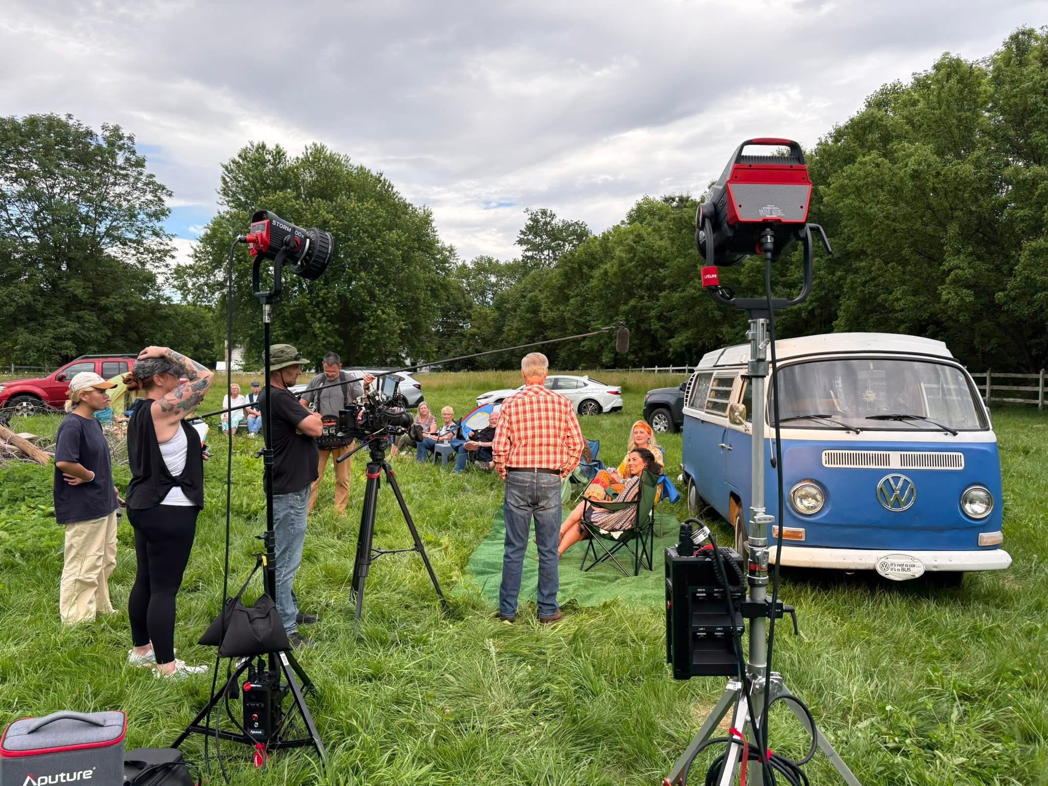A film crew with cameras and lighting equipment filming a vintage Volkswagen van in a grassy field surrounded by trees.