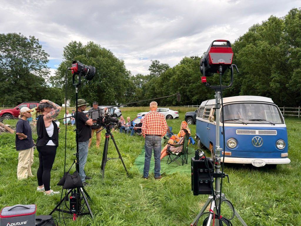 A film crew with cameras and lighting equipment filming a vintage Volkswagen van in a grassy field surrounded by trees.
