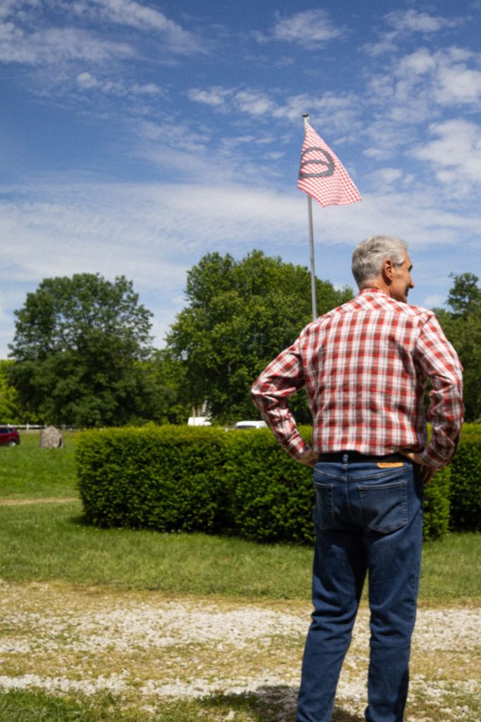 An older man wearing a plaid shirt stands in a grassy field with a red-and-white checkered flag waving in the background.