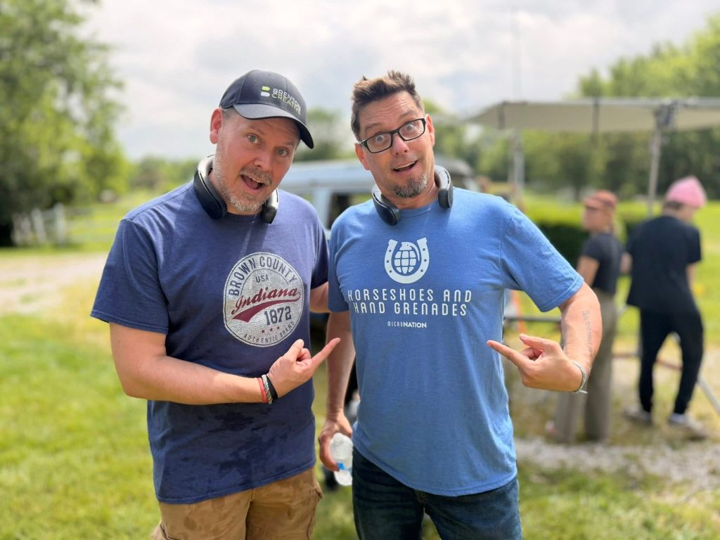 Two men wearing casual clothing and baseball caps, one with a "Recharge" logo and the other with a "Horseshoes and Hand Grenades" logo, standing together and gesturing.