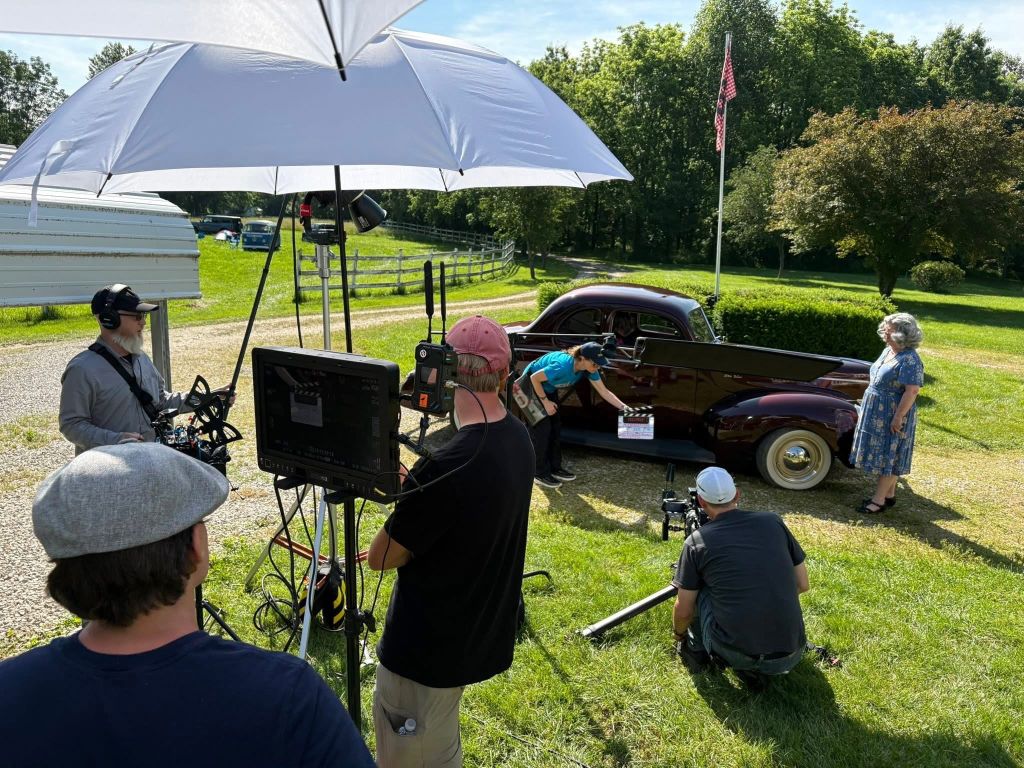 A film crew shooting a scene on a grassy field, with a vintage car and an American flag in the background.
