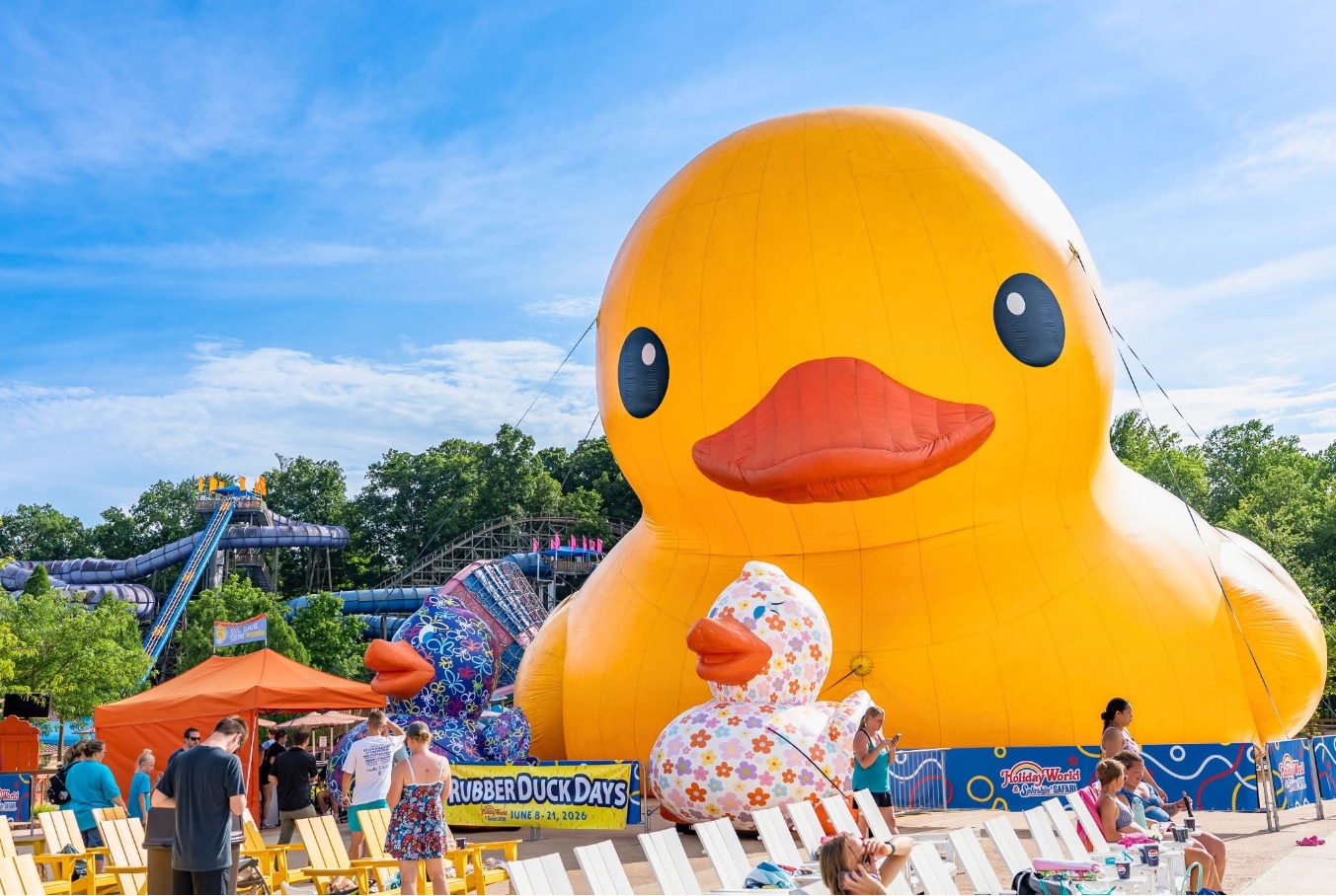 A large inflatable yellow duck with black eyes and an orange beak is the centerpiece of a carnival-like scene, with rides and people in the background.