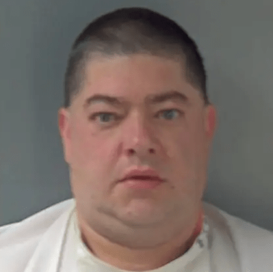 A middle-aged man with a serious expression wearing a white shirt.