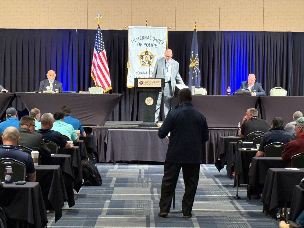 A group of people seated in a conference room, with a Fraternal Order of Police banner and American flag visible. A man is speaking at a podium on the stage.