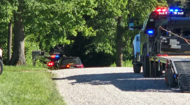Emergency vehicles with flashing lights on a gravel road surrounded by trees.