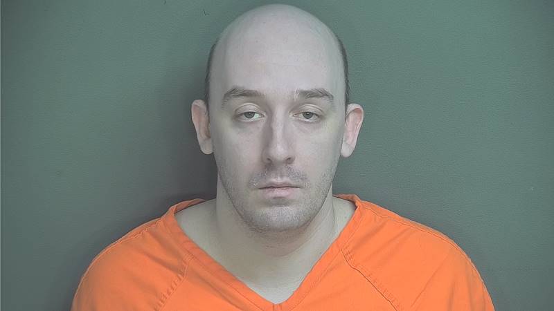 A bald man with a serious expression wearing an orange prison uniform against a green background.