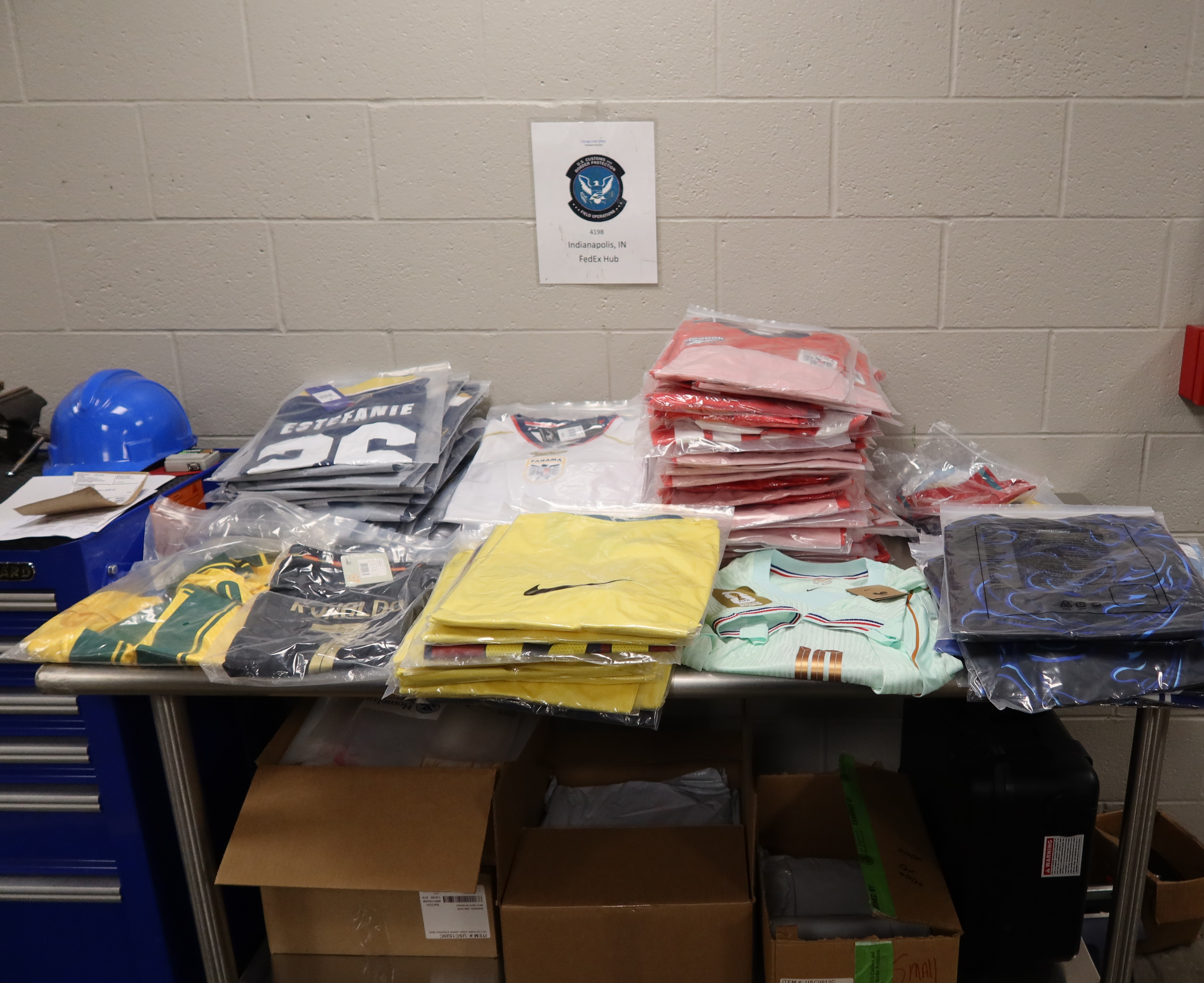 Shelves with various clothing items, including shirts, jackets, and other apparel, in a storage or warehouse setting. A sign on the wall displays "Independence, IN".