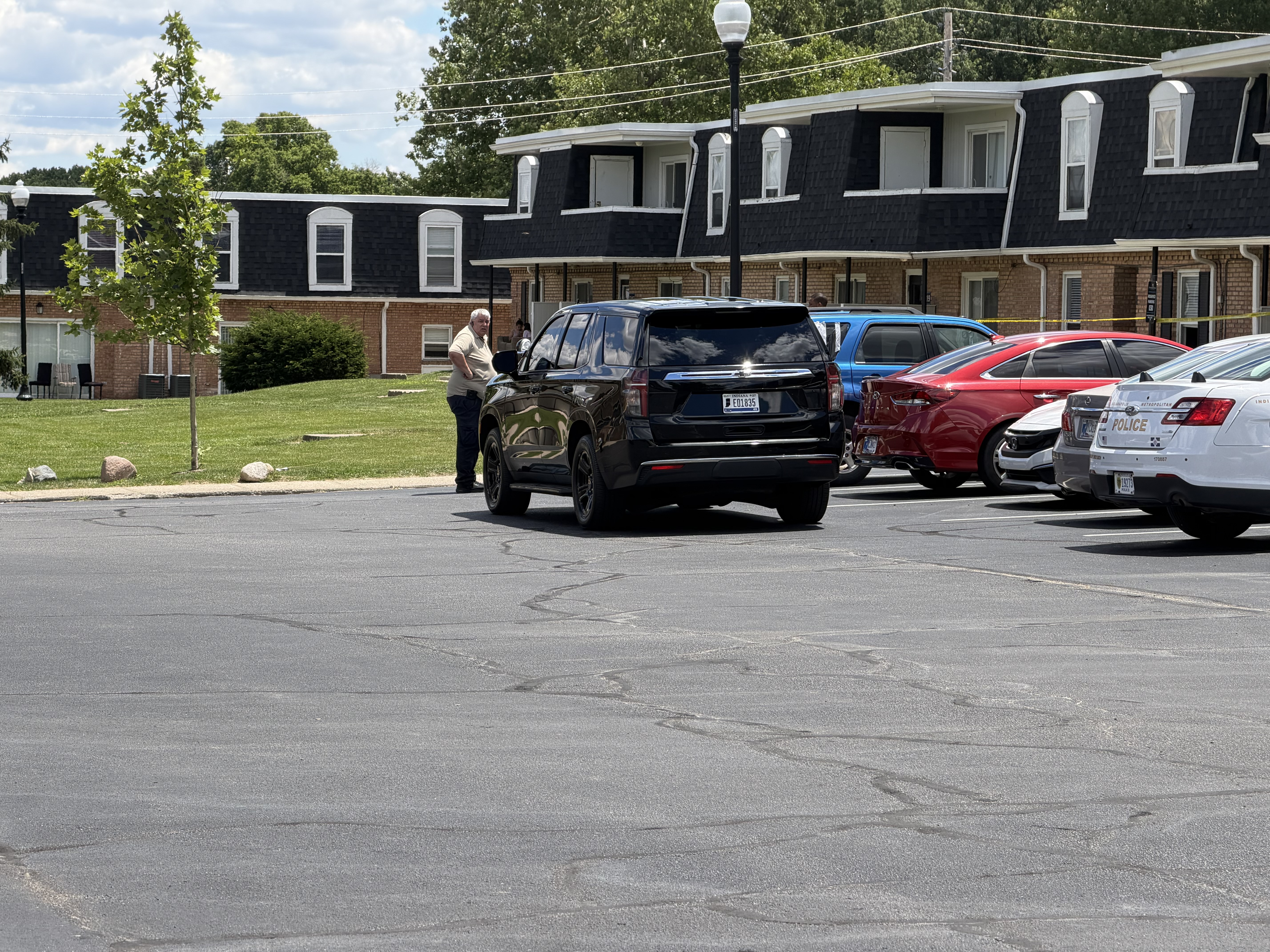 Parking lot with several vehicles, including a black SUV, a red sedan, and a police car. A person is standing near the black SUV.