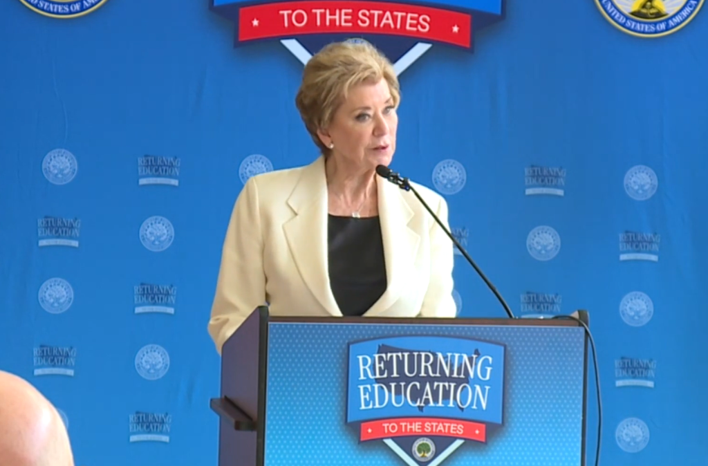 A woman in a cream-colored suit standing at a podium with the "Returning Education to the States" logo, speaking at a public event.
