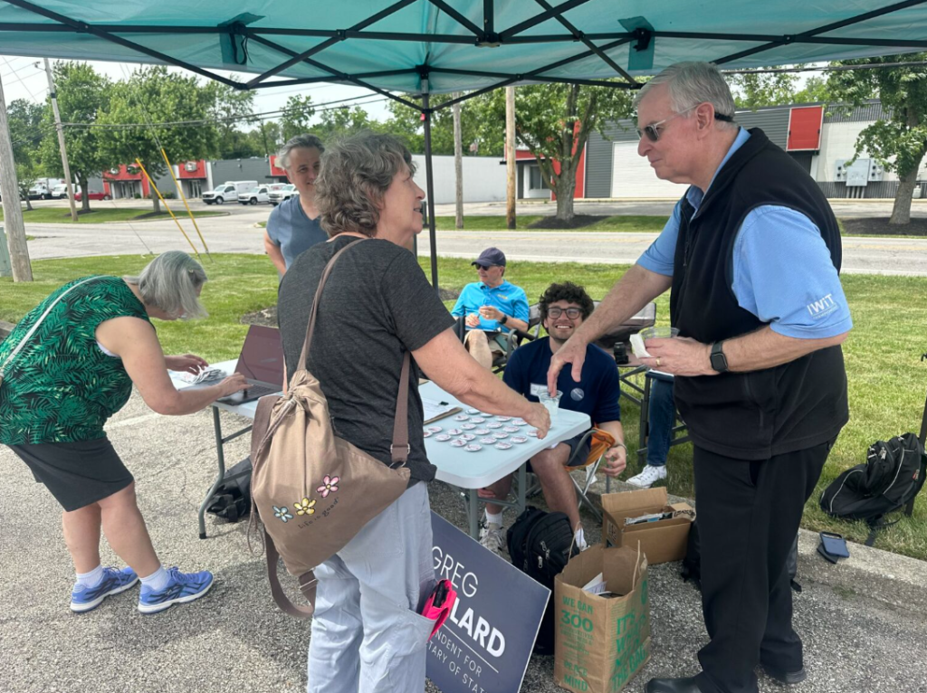 A group of people gathered under a green canopy, some seated at a table with a "Greg Gard" sign, others standing and conversing. Several people appear to be volunteers or organizers.