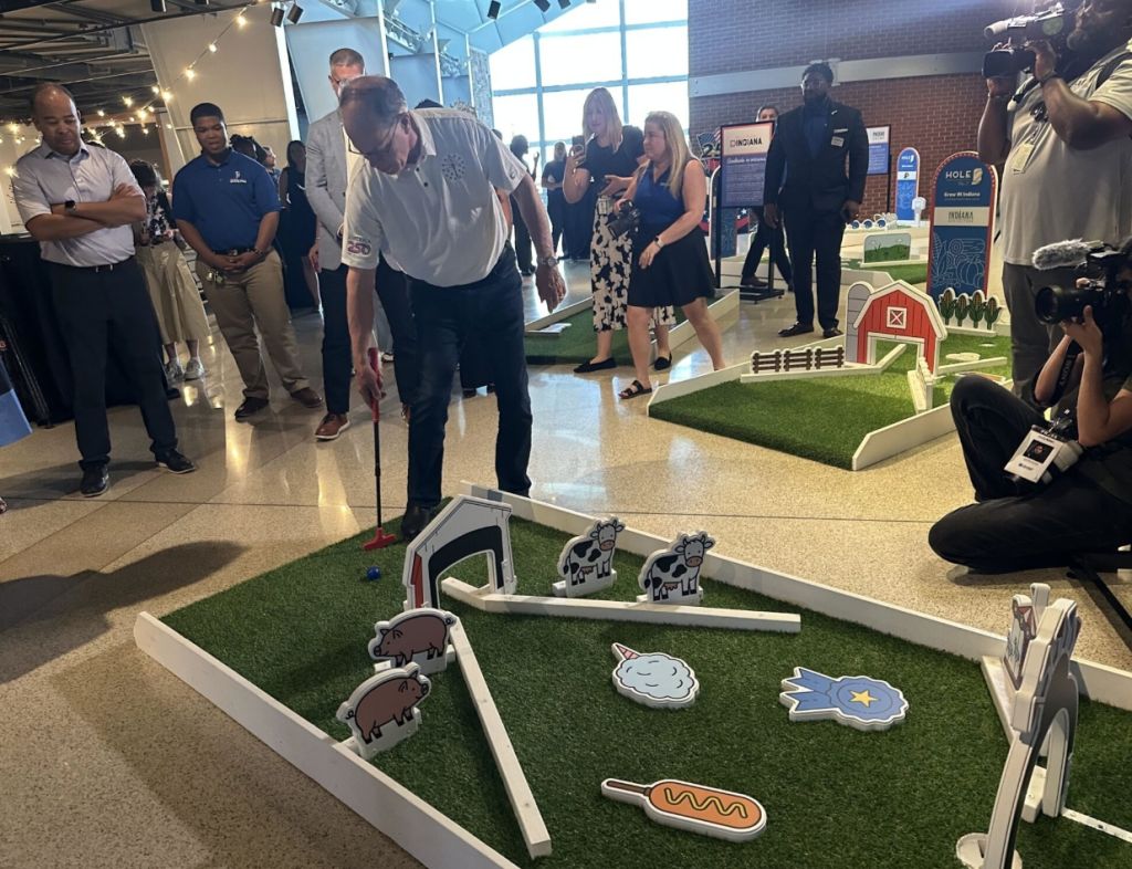 A group of people, including media representatives, gathered around a miniature golf course display with various farm-themed elements like animals and a barn.