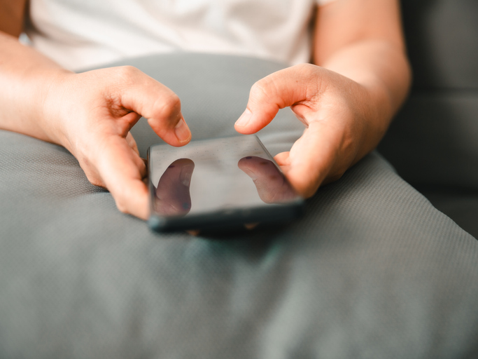 Close-up of a person's hands holding and typing on a smartphone screen while relaxing on a grey couch. Ideal for concepts of mobile communication, social media, technology, and remote work.