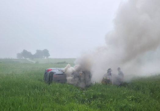 Smoke billows from a damaged vehicle in a grassy field.