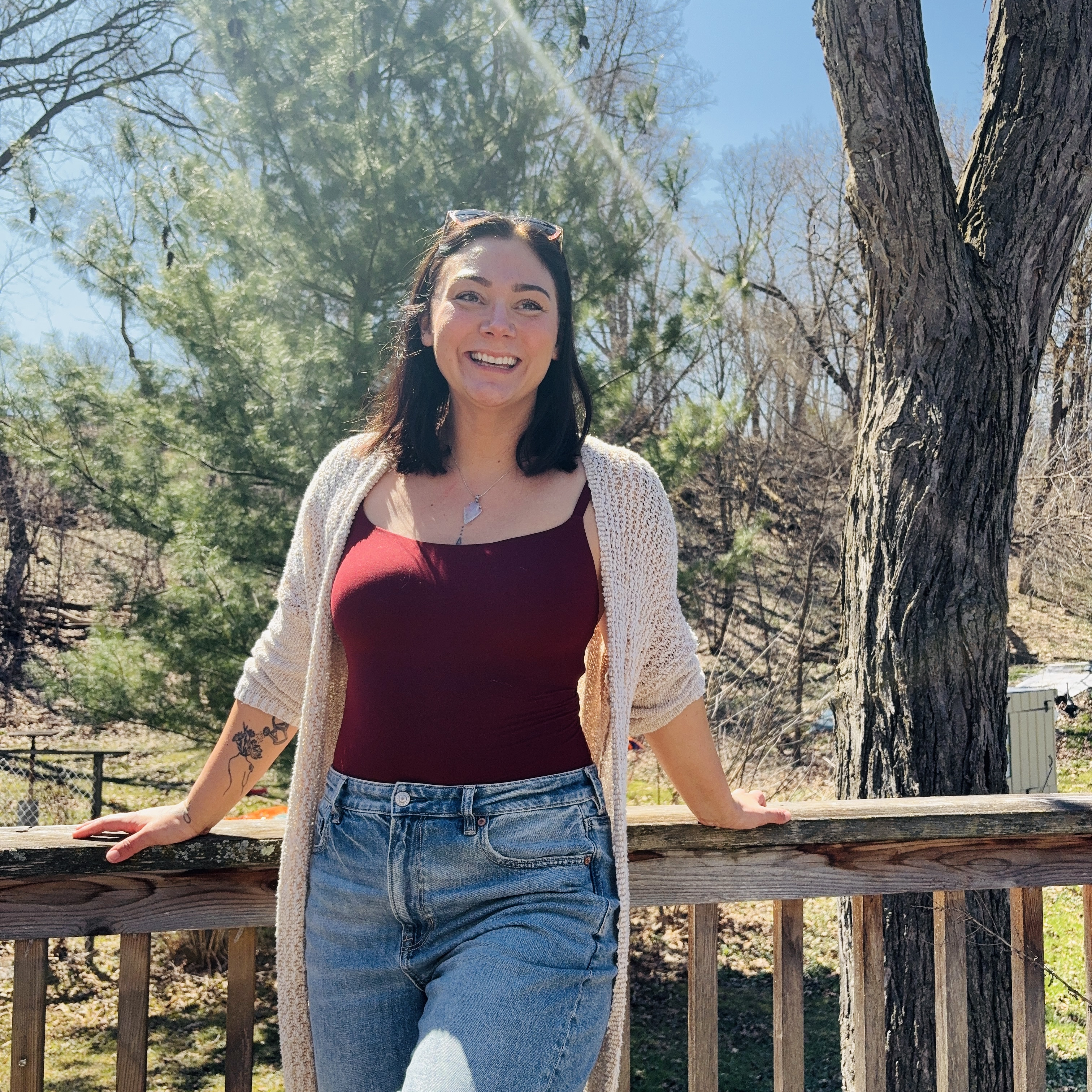 A smiling young woman with long dark hair wearing a red top and a beige cardigan, standing in front of a wooded area.