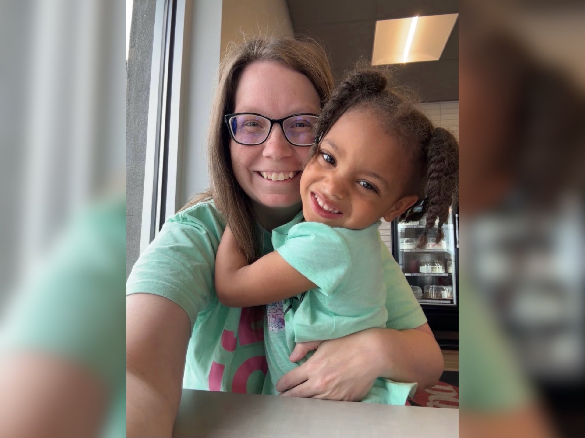 A smiling woman in glasses hugging a young girl with curly hair, both wearing mint green shirts and looking happy.