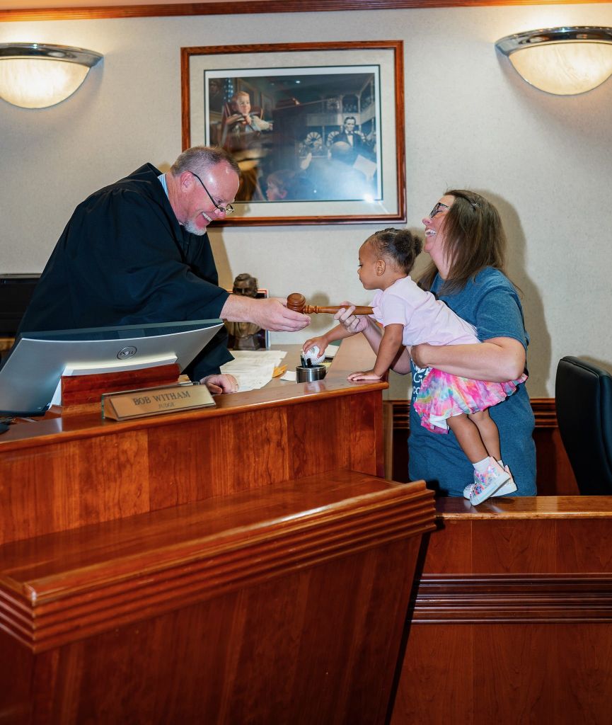 A priest interacting with a young child and woman in a church setting, with a framed photograph on the wall behind them.