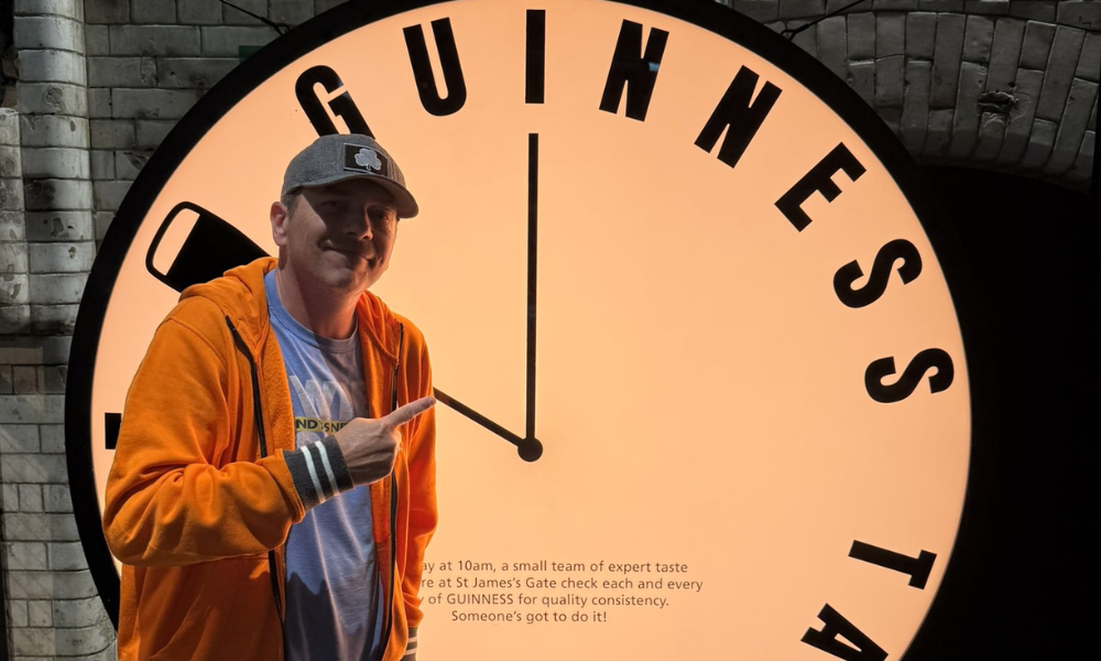 A man in an orange jacket standing in front of a large clock face that says "Guinness's Test 14:00".