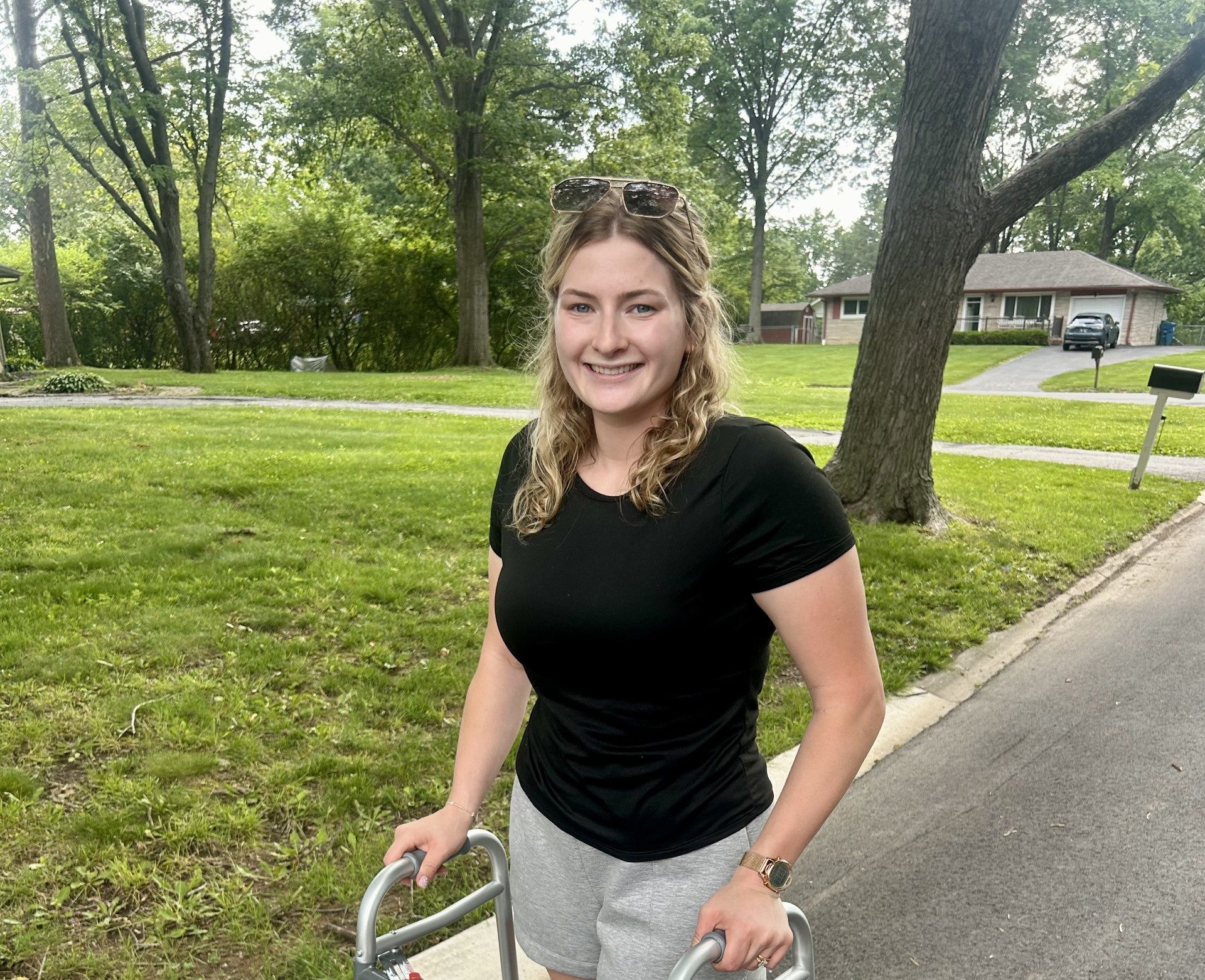 A smiling young woman with curly hair wearing a black shirt and gray shorts, using a walker and standing on a paved path in a grassy, tree-lined neighborhood.